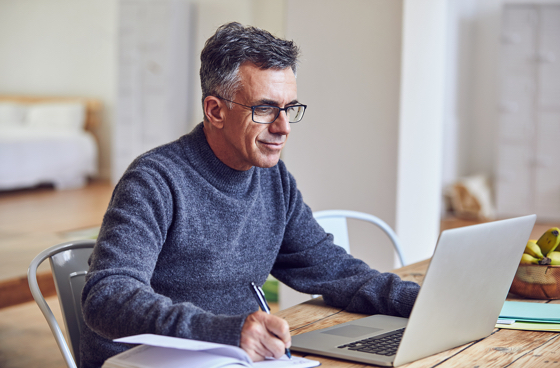 A picture of an older man writing in an notebook while looking at a laptop